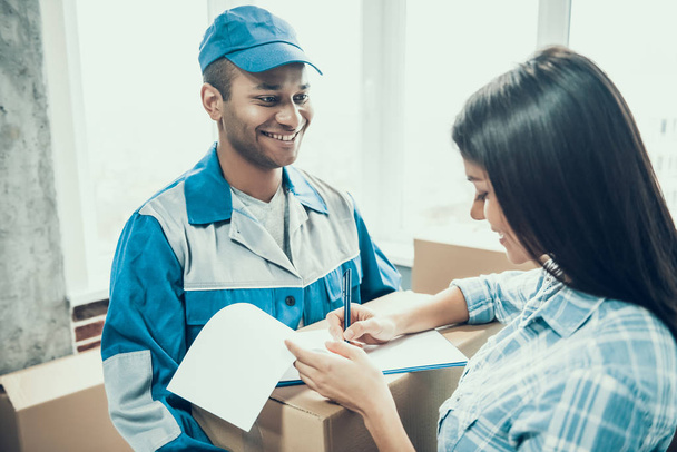 Young Smiling Woman Signing Form for Delivery. Beautiful Girl Receiving Package from Deliveryman in Uniform at Home. Young Man Courier Giving Carton Box to Client in House full of Boxes - Fotografie, Obrázek