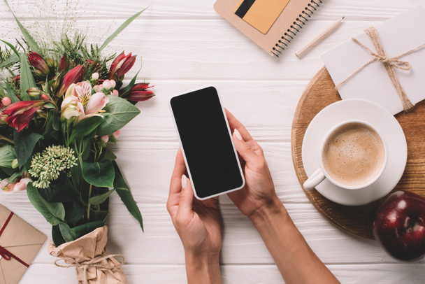 обрізаний знімок жінки, що тримає смартфон з порожнім екраном на поверхні з загорнутим букетом квітів і чашкою кави
 - Фото, зображення