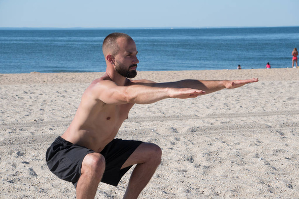 Sportieve jongeman uitoefenen op strand tijdens een warme zomerdag. Squat sit streep opleiding te activeren kern en been spieren bedrijf armen recht naar buiten. - Foto, afbeelding