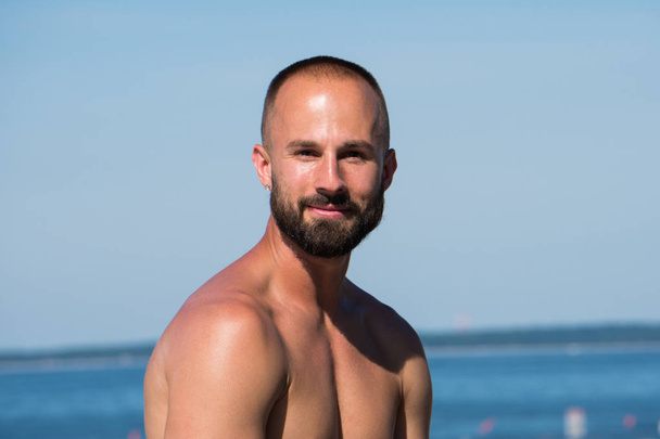 Young handsome athletic adult man with short hair and beard looking into camera while sitting on beautiful tropical beach during hot summer time day. Ocean background scenic view. - Photo, Image