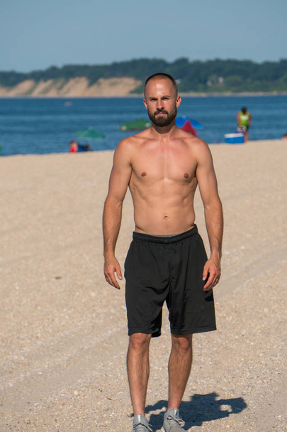 Nuori viehättävä urheilullinen mies seisoo kuumalla kesärannalla päiväsaikaan valmistautuen harjoittelemaan hiekan yli. Ocean tausta ja ihmiset nauttivat sää
 - Valokuva, kuva