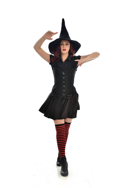 retrato de comprimento total da menina de cabelos vermelhos vestindo traje de bruxa preta e chapéu pontiagudo. em pé pose, isolado no fundo do estúdio branco
. - Foto, Imagem