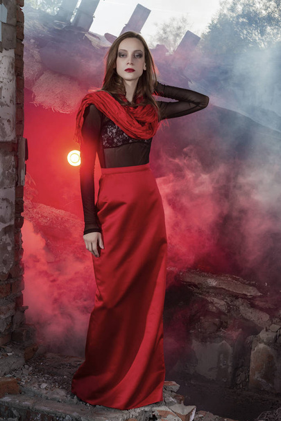 Beautiful fashion model girl wearing a red long skirt and a red shawl poses at the collapsed building, among the ruins of which rises red smoke. Conceptual, fashionable, modern and advertising design. - Foto, Imagem
