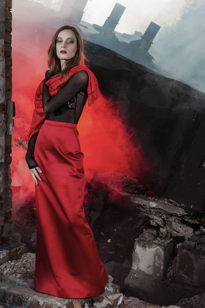 Красивая модель девушки в красной длинной юбке и красной шале позирует у рухнувшего здания, среди руин которого поднимается красный дым. Концептуальный, модный, современный и рекламный дизайн
. - Фото, изображение