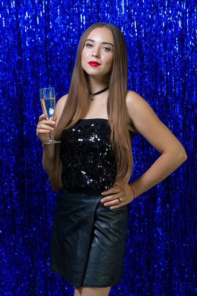 sexy Mädchen mit langen Haaren und rotem Lippenstift auf den Lippen in einem schwarzen Kleid hält ein Glas Champagner auf blauem Hintergrund. - Foto, Bild
