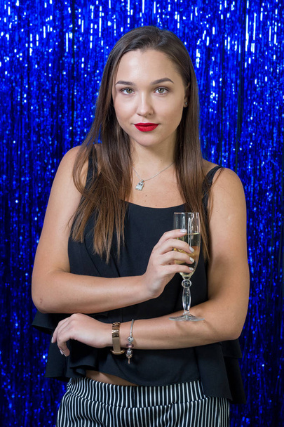 a sexy girl with red lipstick on her lips is holding a glass of champagne in her hands, posing on the camera against the background of shiny blue sequins - Photo, Image