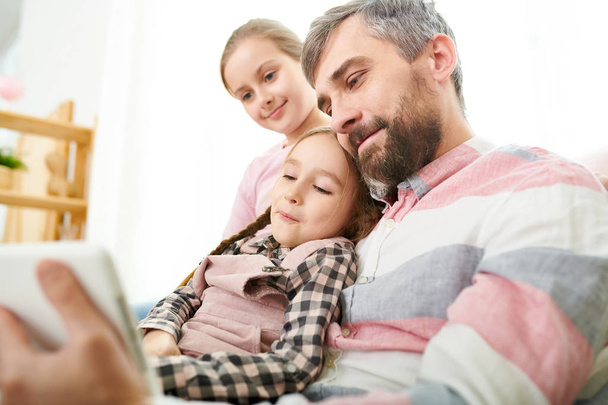 Porträt eines lächelnden reifen Mannes, der ein kleines Mädchen hält, während er Videos auf einem digitalen Tablet mit zwei Töchtern anschaut - Foto, Bild
