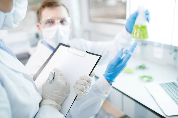 Crop female microbiologist writing down into clipboard test results of research of male holding flask with green solution and pointing at it sitting at desk - Foto, afbeelding