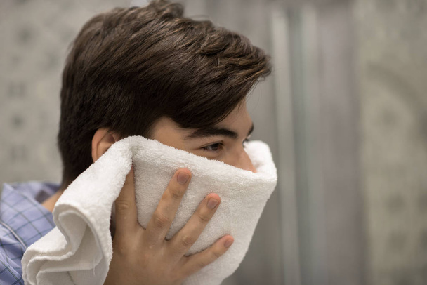 Молодой человек использует белое полотенце, чтобы вытереть лицо во время мытья лица в ванной комнате
.  - Фото, изображение