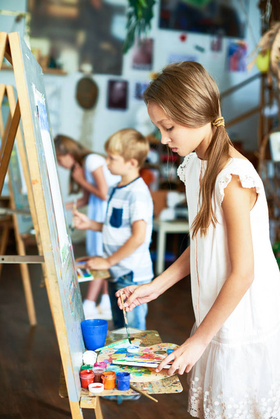 Side view portrait of pretty girl painting picture on easel in art class with other children in background - Photo, Image