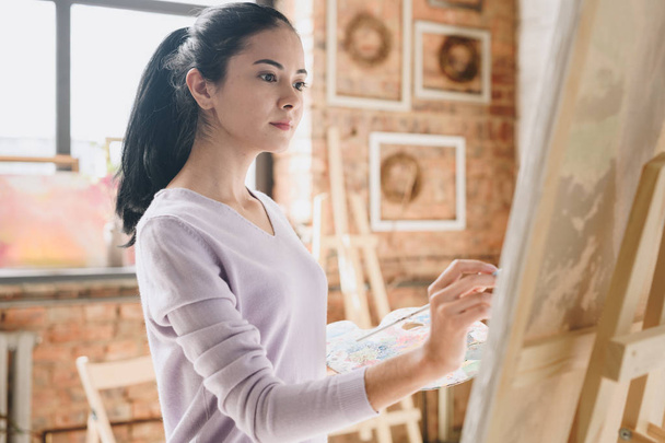 Waist up portrait of inspired young woman holding palette painting pictures at easel in art studio standing against windows in sunlight - 写真・画像
