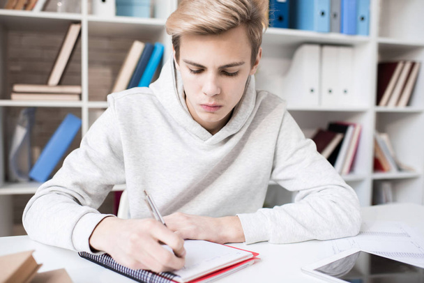 Young concentrated Caucasian male student writing notes in copybook while sitting at desk in classroom - Valokuva, kuva