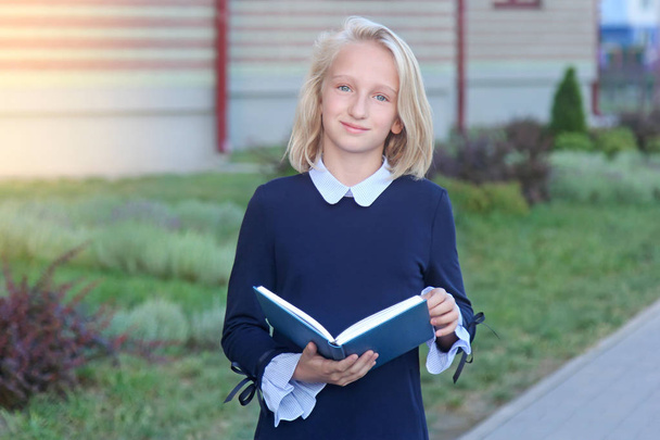Beautiful blonde girl in elegant dress with book in hands near school. Schoolgirl likes to learn and read. - Zdjęcie, obraz
