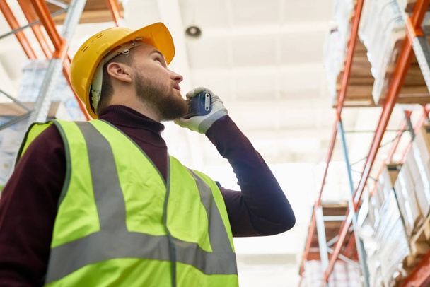 Low angle portrait of young man wearing hardhat and reflective jacket speaking by walkie-talkie looking up tall shelves in warehouse, copy space - Foto, imagen