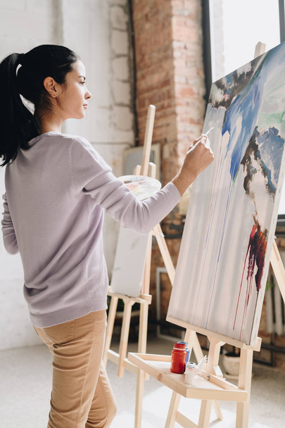 Po stronie widoku portret młodego artysty kobiet posiadających palety malarstwo obrazy sztalugowe w stojący studio sztuki w słońcu - Zdjęcie, obraz