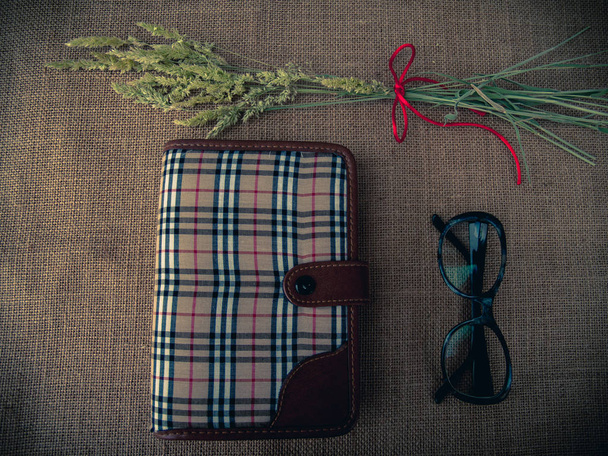 Vintage style. Organized desk with closed diary, eye glasses, dry grass and burlap background - Photo, Image