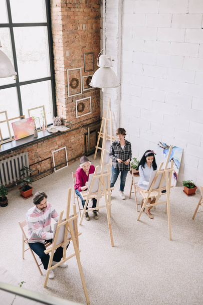 Above view portrait of adult students painting sitting by easels in art studio with female art teacher watching them in spacious sunlit loft space - Photo, Image