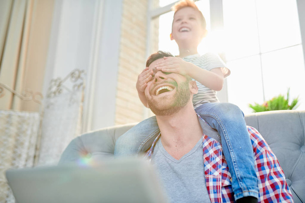 Милый маленький мальчик с широкой улыбкой сидит на плечах своего бородатого отца и закрывает глаза руками, веселясь вместе дома, блики линзы
 - Фото, изображение