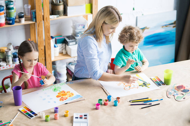 Retrato en ángulo alto de una joven dibujando con dos niños sentados en una mesa de madera en un estudio de arte moderno para niños
 - Foto, imagen