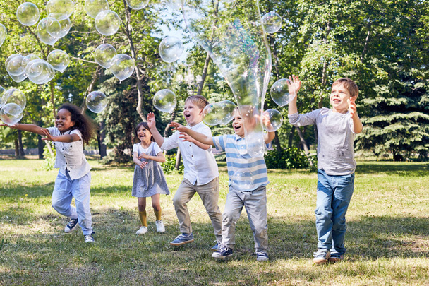 Groupe multi-ethnique d'enfants joyeux attrapant de grosses bulles de savon tout en faisant la fête au parc public vert illuminé avec des rayons de soleil
 - Photo, image