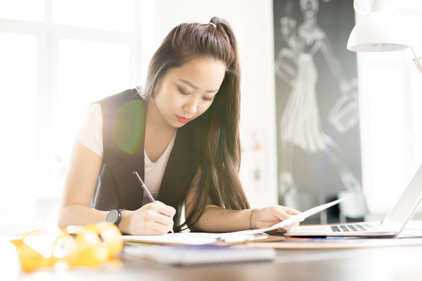 Porträt einer kreativen jungen asiatischen Frau, die Skizzen für maßgeschneiderte Kleidung zeichnet, während sie am Arbeitstisch im sonnenbeschienenen Modedesign-Atelier steht, Linsenschlag - Foto, Bild