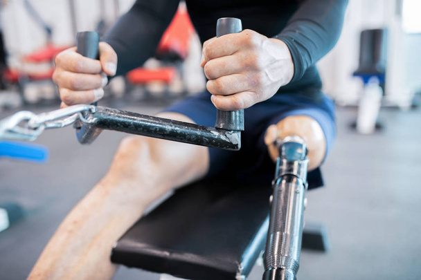 Keskiosa lähikuva tunnistamaton lihaksikas mies proteesi jalka käyttäen painokoneita treenatessa kuntosalilla
 - Valokuva, kuva
