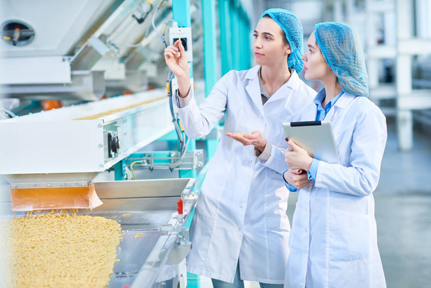 Portret van twee jonge vrouwelijke werknemers dragen Labjassen permanent door transportband lijn met macaroni inspectie van kwaliteit in schone productie werkplaats, kopie ruimte - Foto, afbeelding