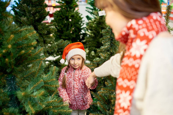 Высокоугловой портрет милой маленькой девочки в шляпе Санты, держащей за руку матерей, выбирая елку на рынке в торговом центре
 - Фото, изображение