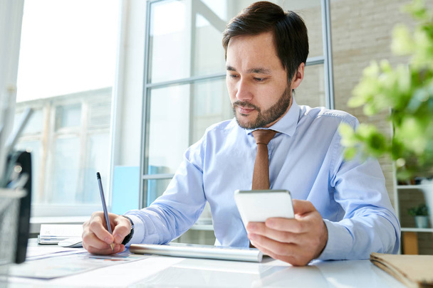 Stylish bearded man using smartphone and taking notes on paper working at table in office.  - Photo, Image