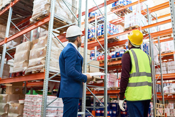 Back view portrait of warehouse manager and worker in hardhats doing stock inventory in warehouse, looking up at tall shelves with goods - Photo, Image