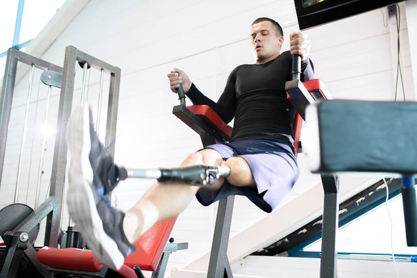 Niedrigwinkel-Porträt eines gut aussehenden, muskulösen Mannes mit Beinprothese, der Klimmzüge macht, während er im modernen Fitnessstudio trainiert - Foto, Bild
