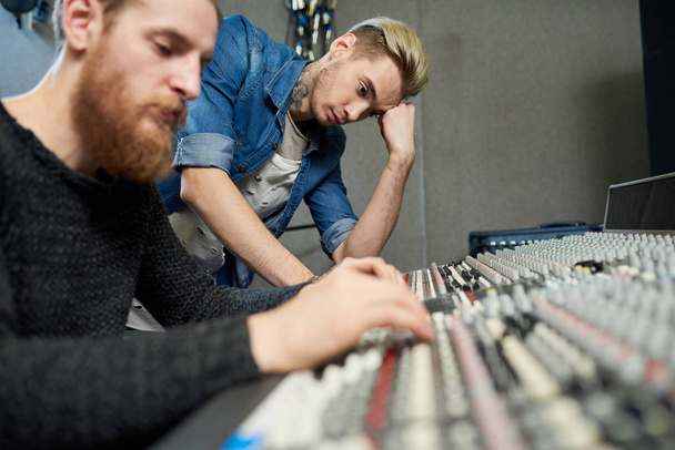 Jonge mannen permanent op elektronische console in studio en bezig met muziek maken.  - Foto, afbeelding