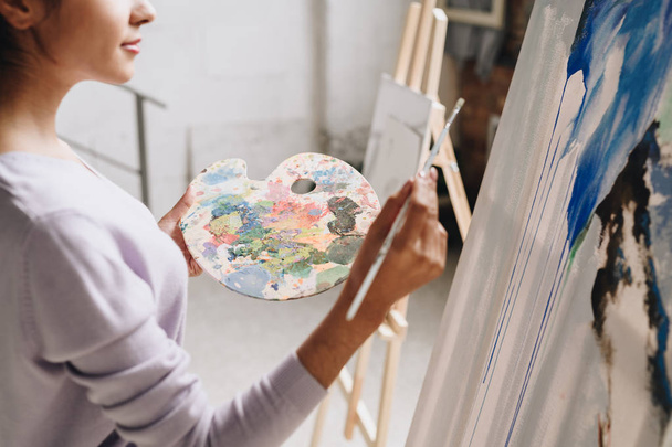 Mid-section portrait of young female artist holding palette painting pictures at easel in art studio standing against windows in sunlight - Foto, Imagem