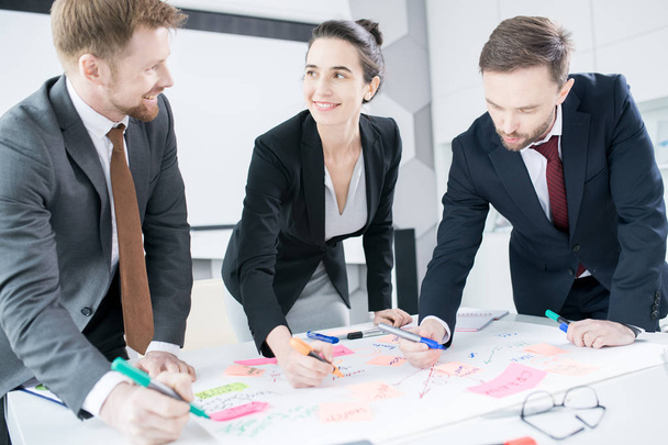 Portrait of three businesspeople with smiling young woman  standing at table in conference room  drawing charts while planning strategy, copy space - Photo, Image