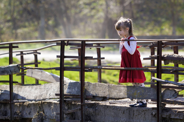 Melko pieni vaalea pitkäkarvainen tyttö mukava punainen mekko seisoo yksin vanha sementti silta nojaten puiset kaiteet etsivät tarkkaavaisesti alas lämmin aurinkoinen päivä. Lapset uteliaisuus ja fantasiat käsite
. - Valokuva, kuva