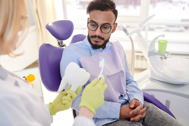 Odontólogo rubio irreconocible que explica al joven paciente mestizo cómo cepillarse los dientes correctamente mientras tiene cita en la oficina moderna
 - Foto, imagen