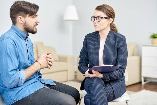 Handsome bearded man suffering from depression having consultation with highly professional psychiatrist at cozy office, she listening to him with concentration - Photo, Image