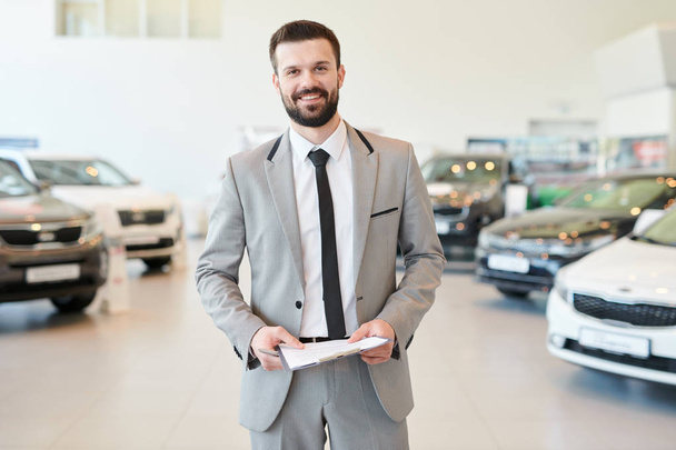 Portrait of smiling bearded salesman holding contract while standing at auto salon - Foto, Imagen