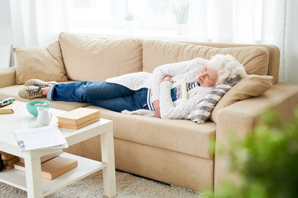 Грустная депрессивная красивая пожилая женщина в повседневной одежде лежит на диване и обнимает фото с мужем во время сна в гостиной
 - Фото, изображение
