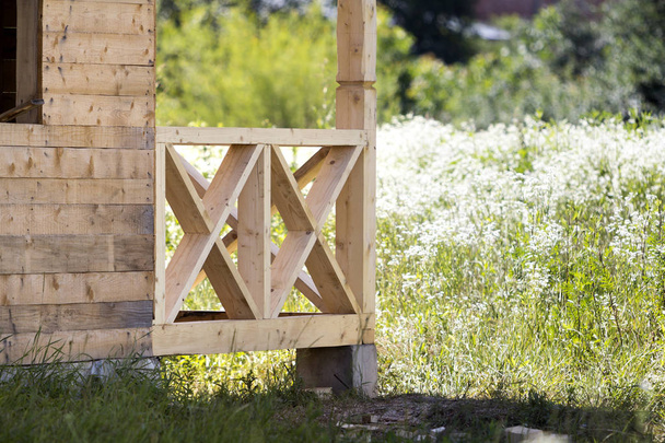 Close-up section geometrical decorative simple detail of new natural not painted wooden planks and bars for balcony, attic fence on blurred blooming summer field background. Construction and design. - Photo, Image