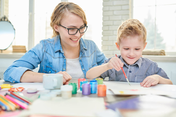 Ritratto di felice giovane madre che dipinge con il suo simpatico figlio seduto al tavolo da disegno insieme contro grandi finestre nella scuola di sviluppo
 - Foto, immagini