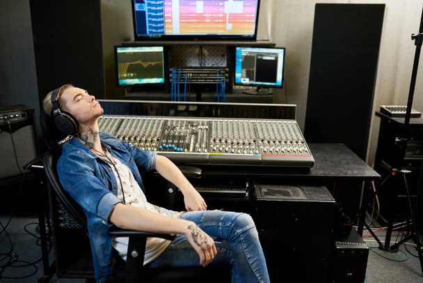 Side view of stylish man in headphones listening to music with enjoyment sitting at console in recording studio.  - Фото, изображение