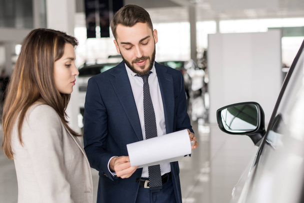 Taille portret van knappe autoverkoper praten met jonge vrouw helpt haar kiezen in auto showroom - Foto, afbeelding