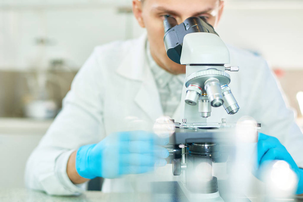Працьовитий молодий вчений в гумових рукавичках і білому пальто дивиться через мікроскоп під час роботи над створенням штучного м'яса в сучасній лабораторії
 - Фото, зображення