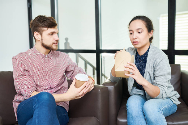 Nuori valkoihoinen parrakas mies puhuu naispuoliselle kollegalleen, kun hän syö noutoruokaa toimistossa taukohuoneessa
 - Valokuva, kuva