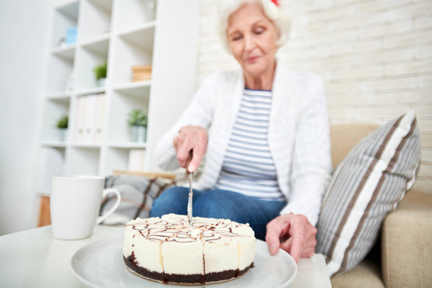 Calme belle vieille femme coupe gâteau avec couteau tout en se préparant à boire du thé seul sur son anniversaire à la maison
 - Photo, image