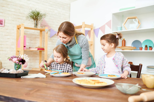 Mooie jonge vrouw dragen schort en haar schattige kleine dochters hebben van pret terwijl het verfraaien van smakelijk gebak met kleurrijke glazuur, interieur van moderne eetkamer op achtergrond - Foto, afbeelding