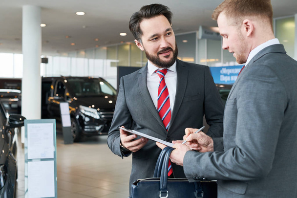 Businessman standing at auto salon with salesman. He is signing a contract - Foto, Imagem