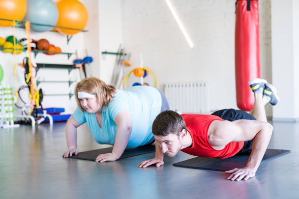 Retrato de cuerpo entero del entrenador personal guapo haciendo ejercicios de fitness en el gimnasio con mujer con sobrepeso en segundo plano
 - Foto, Imagen