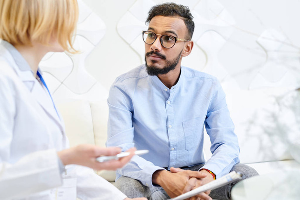 Portrait shot of mixed-race bearded patient sitting on cozy sofa and talking to female physician while having consultation at modern office - Photo, Image